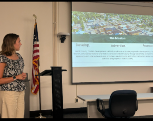 A young woman (Emily) in a flower blouse and slacks is standing in a conference room to the left side of a slide show presentation showing an overhead view of a town on top and text below.